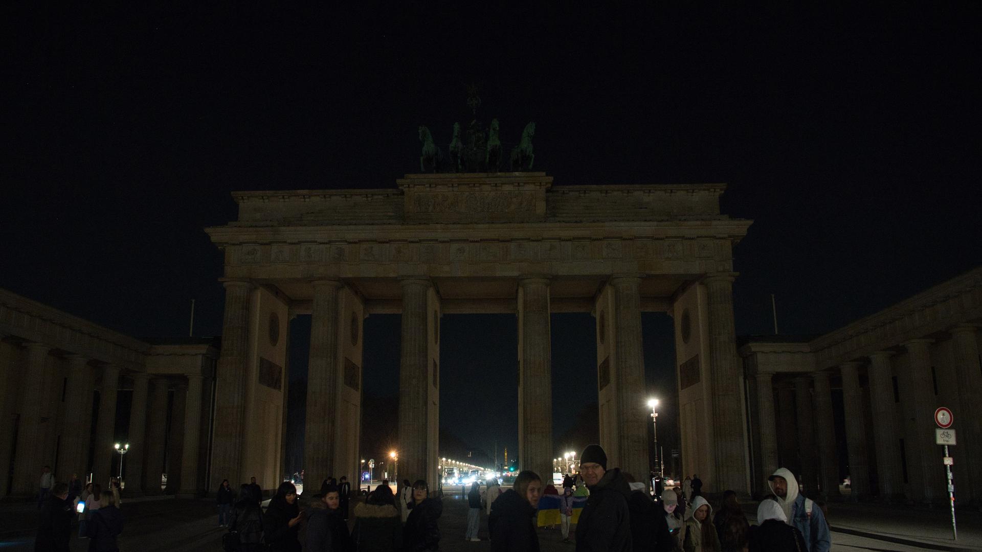 Berlin: Menschen stehen vor dem dunklen Brandenburger Tor. Für eine Stunde wurde die Beleuchtung ausgemacht. 