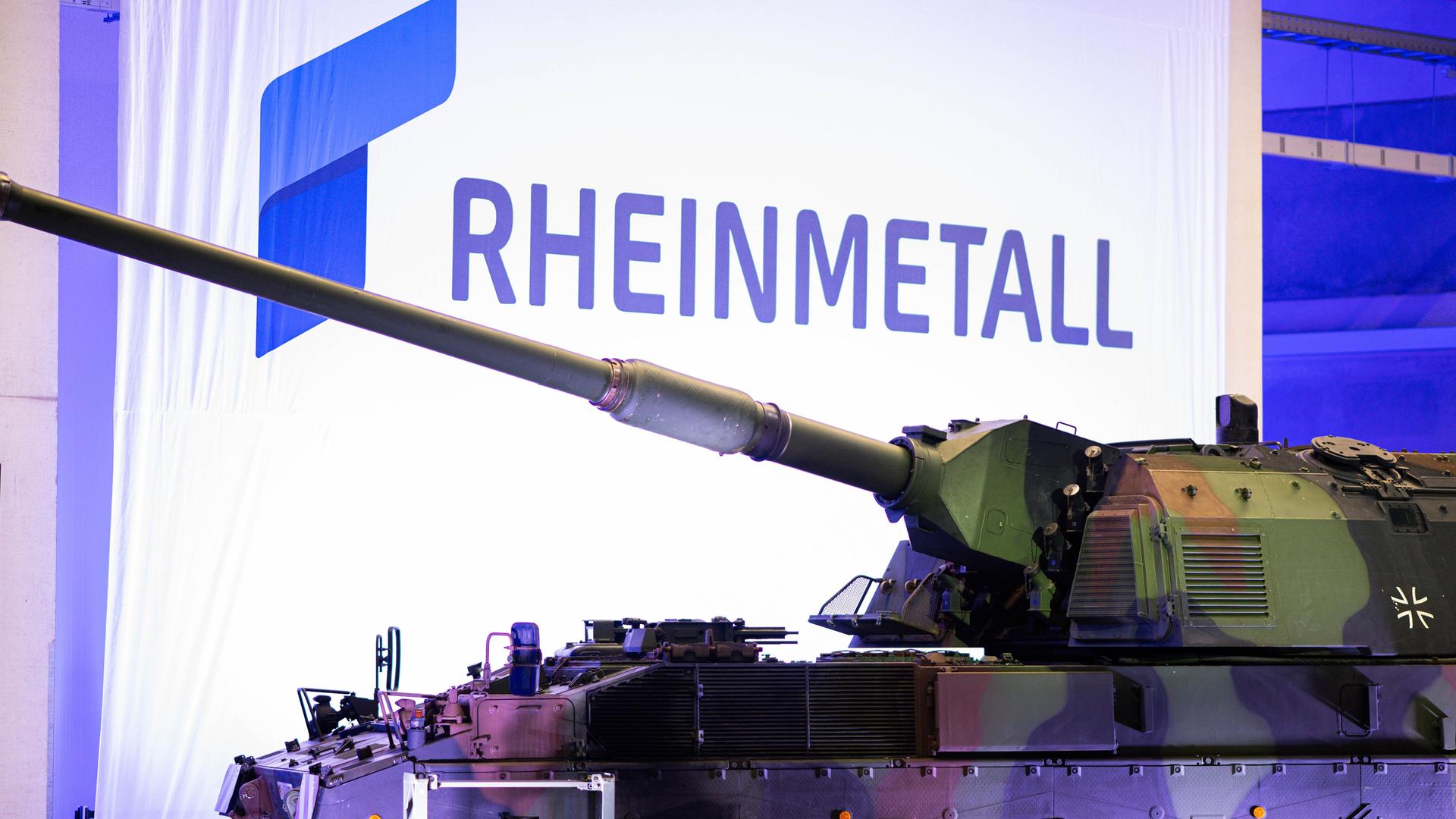 Zu sehen ist ein Panzer in Tarnfarben, der in einer Produktionshalle steht. Im Hintergrund: Das Wort „RHEINMETALL“ (geschrieben in blauben Großbuchstaben) Zu sehen ist ein Panzer in Tarnfarben, der in einer Produktionshalle steht. Im Hintergrund: Das Wort „RHEINMETALL“ (geschrieben in blauben Großbuchstaben)