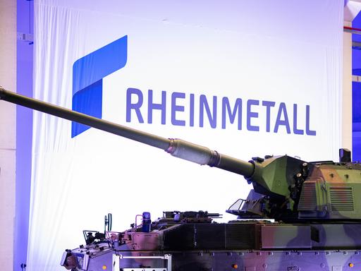 Zu sehen ist ein Panzer in Tarnfarben, der in einer Produktionshalle steht. Im Hintergrund: Das Wort „RHEINMETALL“ (geschrieben in blauben Großbuchstaben) Zu sehen ist ein Panzer in Tarnfarben, der in einer Produktionshalle steht. Im Hintergrund: Das Wort „RHEINMETALL“ (geschrieben in blauben Großbuchstaben)