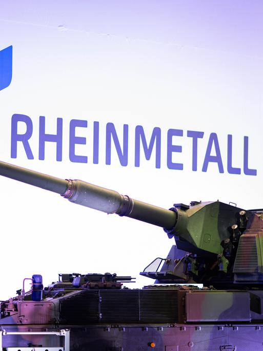 Zu sehen ist ein Panzer in Tarnfarben, der in einer Produktionshalle steht. Im Hintergrund: Das Wort „RHEINMETALL“ (geschrieben in blauben Großbuchstaben) Zu sehen ist ein Panzer in Tarnfarben, der in einer Produktionshalle steht. Im Hintergrund: Das Wort „RHEINMETALL“ (geschrieben in blauben Großbuchstaben)