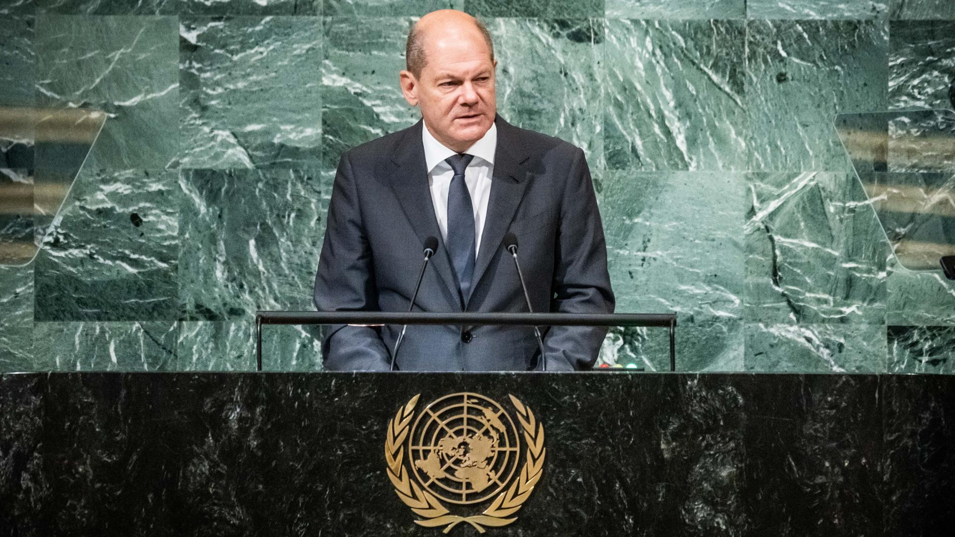 Bundeskanzler Olaf Scholz steht an einem Rednerpult, auf der Trennwand zum Publikum ist das goldene Wappen der Vereinten Nationen zu sehen.
