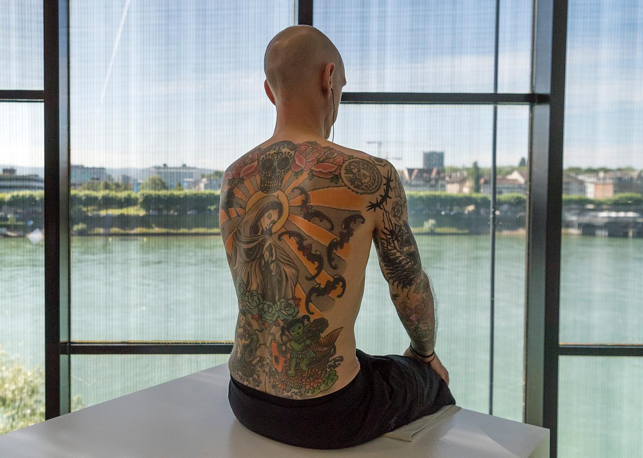 Tim Steiner, das Vorbild des Filmes "Der Mann, der seine Haut verkaufte", bei einer Ausstellung im Museum Tinguely in Basel. Wir blicken auf seinen bunt tätowierten Rücken, im Hintergrund ist durch die Museumsscheibe Wasser zu sehen. 