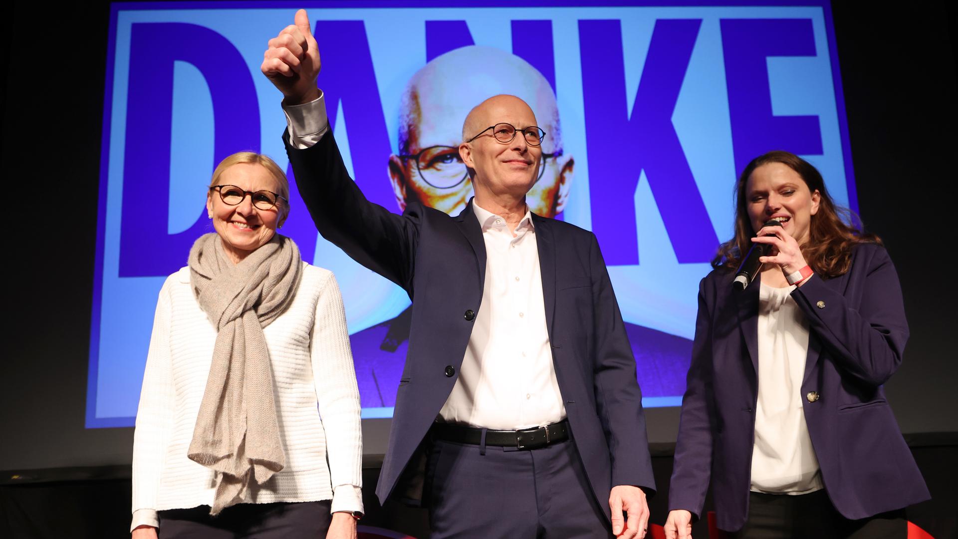 Peter Tschentscher steht mit Parteifreundinnen auf einer Bühne vor SPD-Anhängern. Mit seiner rechten Hand zeigt er ein siegreiches "Daumen-hoch-Symbol". Im Hintergrund ist eine Projektion seines Gesichtes mit einen großen "Danke"-Schriftzug zu sehen. 