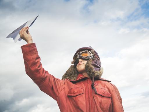Ein Junge in Pilotenkostüm hält einen Papierflieger in die Luft.