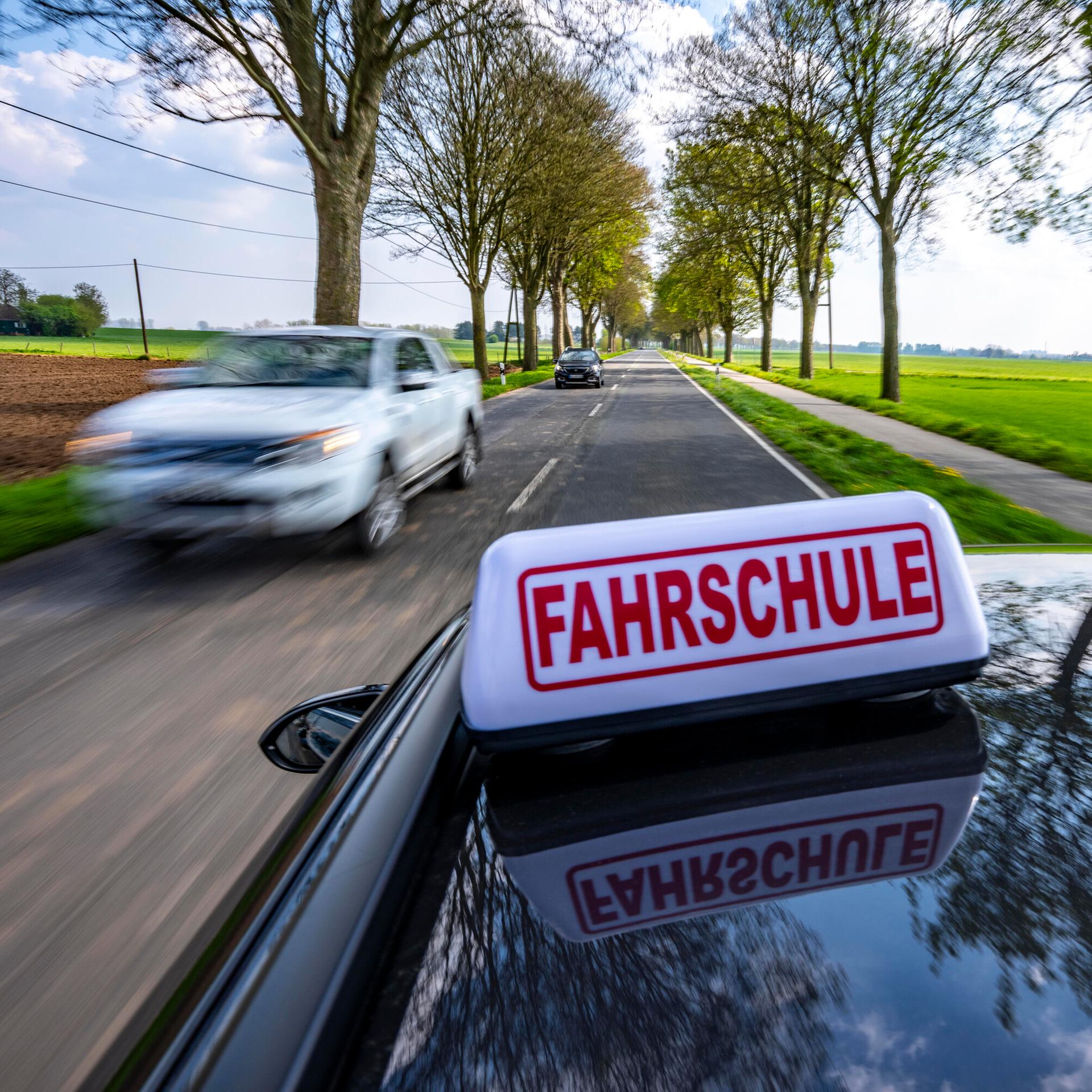 Ein schwarze Auto mit einem Schild "Fahrschule" auf dem Dach fährt auf einer Landstraße entlang