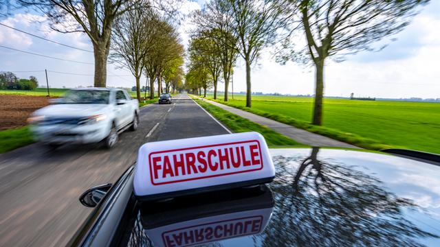 Ein schwarzes Auto mit einem Schild "Fahrschule" auf dem Dach fährt auf einer Landstraße entlang.