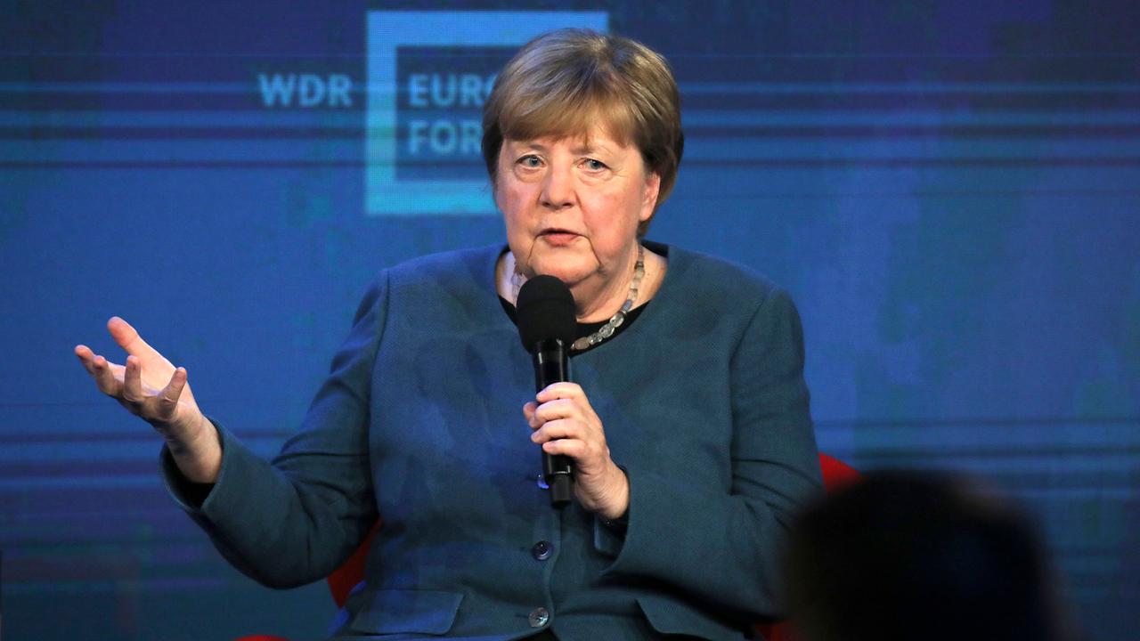 Merkel sitzt auf einem Podium und hält ein Mikrofon in der Hand. Mit der anderen Hand gestikuliert sie.