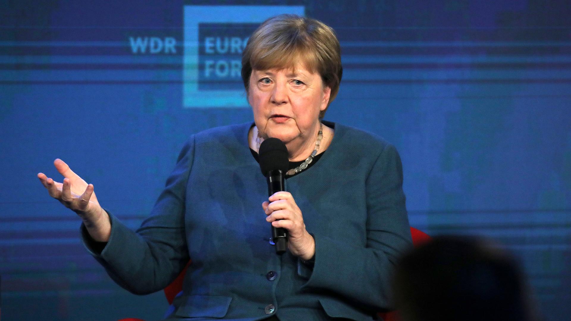 Merkel sitzt auf einem Podium und hält ein Mikrofon in der Hand. Mit der anderen Hand gestikuliert sie.
