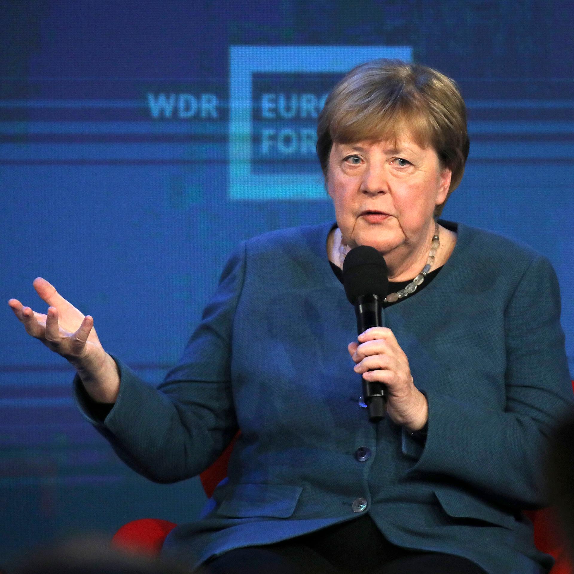 Merkel sitzt auf einem Podium und hält ein Mikrofon in der Hand. Mit der anderen Hand gestikuliert sie.