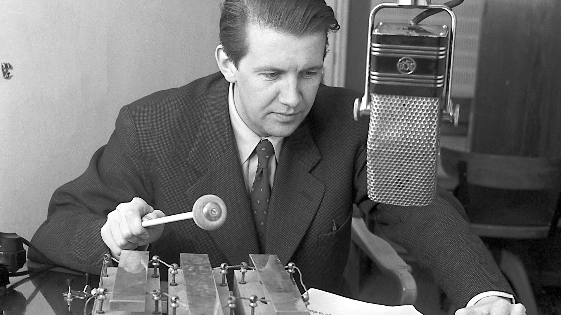 Der schwedische Radiojournalist Folke Olhagen sitzt 1953 hinter dem Mikfrofon eines Radiostudios und spielt dabei mit einem Schlagstock auf drei großen Glockenspiel-Tönen