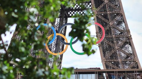 Am Eiffelturm hängen die Olympischen Ringen. Sie sind aus der Ferne leicht durch Baumblätter verdeckt.