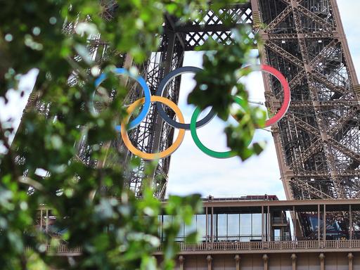 Am Eiffelturm hängen die Olympischen Ringen. Sie sind aus der Ferne leicht durch Baumblätter verdeckt.