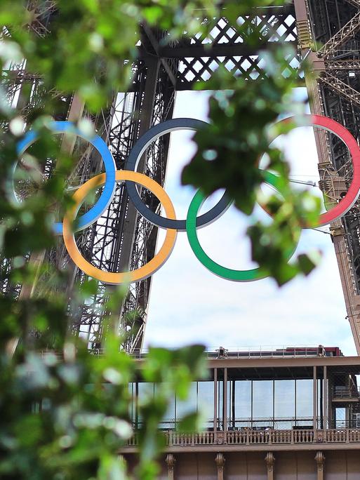 Am Eiffelturm hängen die Olympischen Ringen. Sie sind aus der Ferne leicht durch Baumblätter verdeckt.