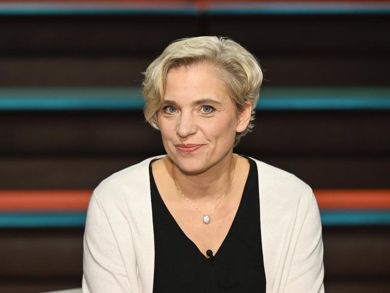 Bente Scheller trägt ein schwarzes Oberteil, eine helle Strickjacke und schaut in einem Fernsehstudio freundlich in die Kamera