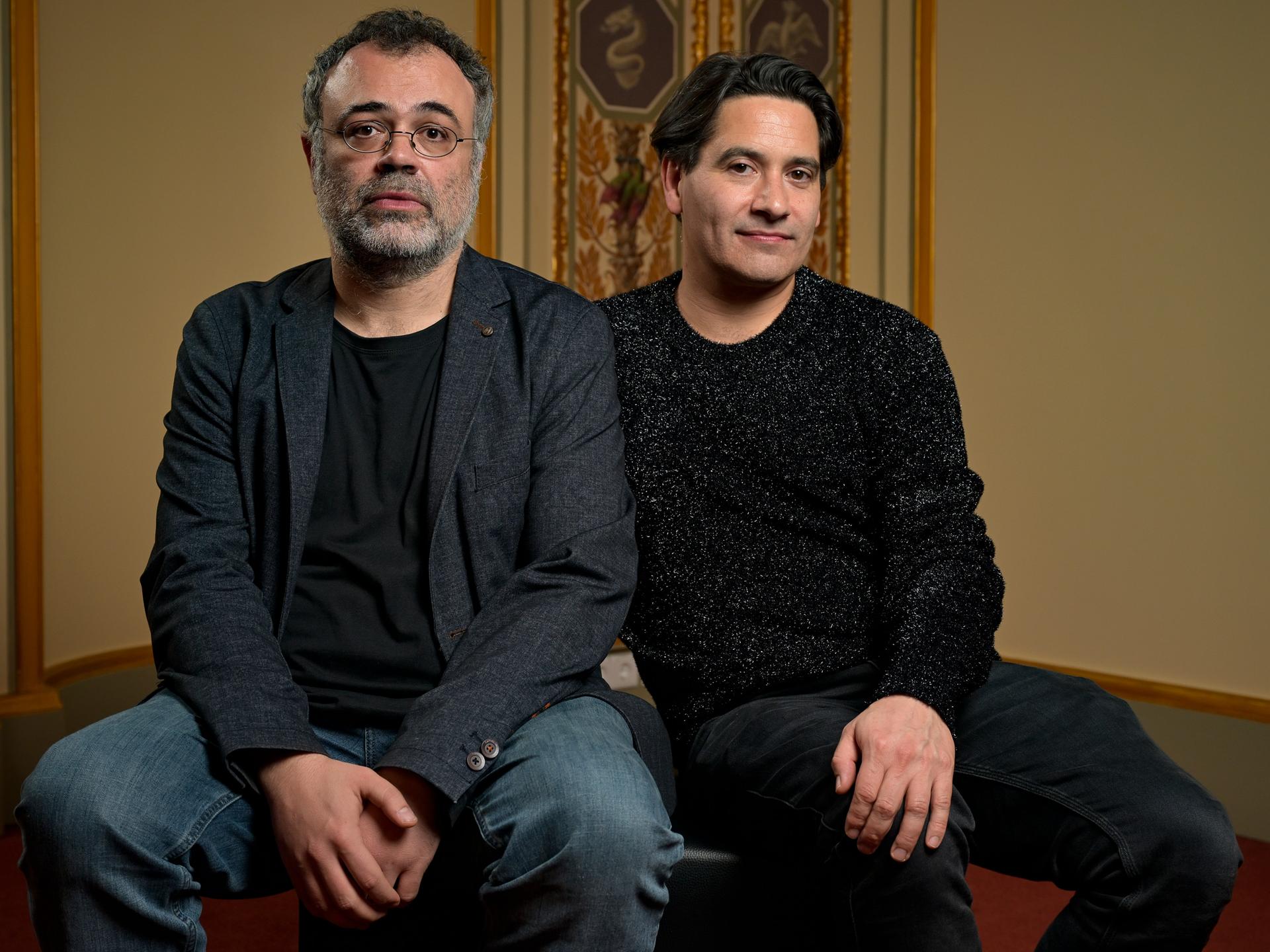 Zwei Männer sitzen auf einem Hocker vor historisch wirkender Fassade. Beide sind dunkel gekleidet und schauen ernst bis neugierig. Der linke hat eine runde Brille und einen grau-schwarzen Bart, der rechte dunkles Haar. 