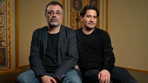 Zwei Männer sitzen auf einem Hocker vor historisch wirkender Fassade. Beide sind dunkel gekleidet und schauen ernst bis neugierig. Der linke hat eine runde Brille und einen grau-schwarzen Bart, der rechte dunkles Haar. 