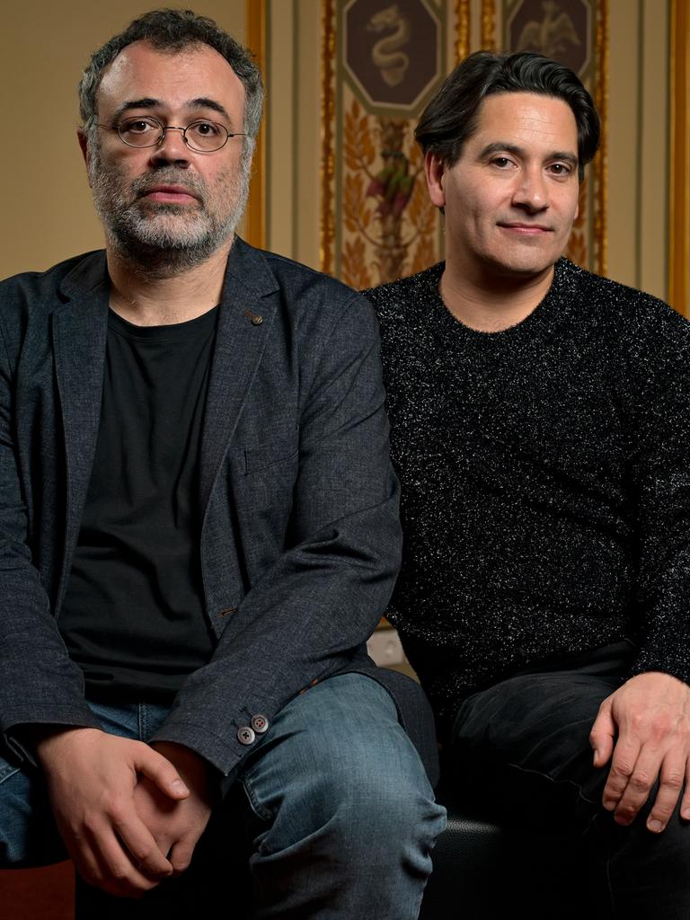 Zwei Männer sitzen auf einem Hocker vor historisch wirkender Fassade. Beide sind dunkel gekleidet und schauen ernst bis neugierig. Der linke hat eine runde Brille und einen grau-schwarzen Bart, der rechte dunkles Haar. 