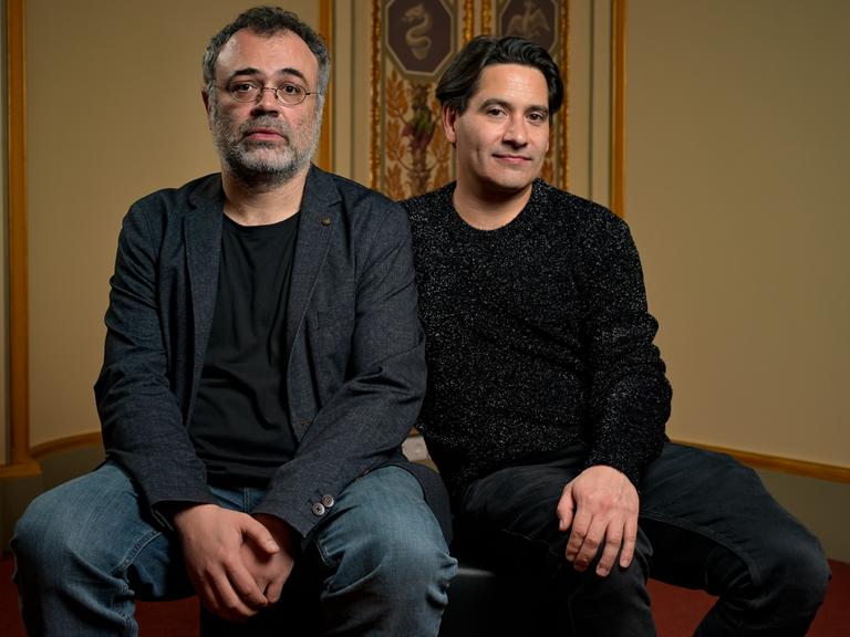 Zwei Männer sitzen auf einem Hocker vor historisch wirkender Fassade. Beide sind dunkel gekleidet und schauen ernst bis neugierig. Der linke hat eine runde Brille und einen grau-schwarzen Bart, der rechte dunkles Haar. 