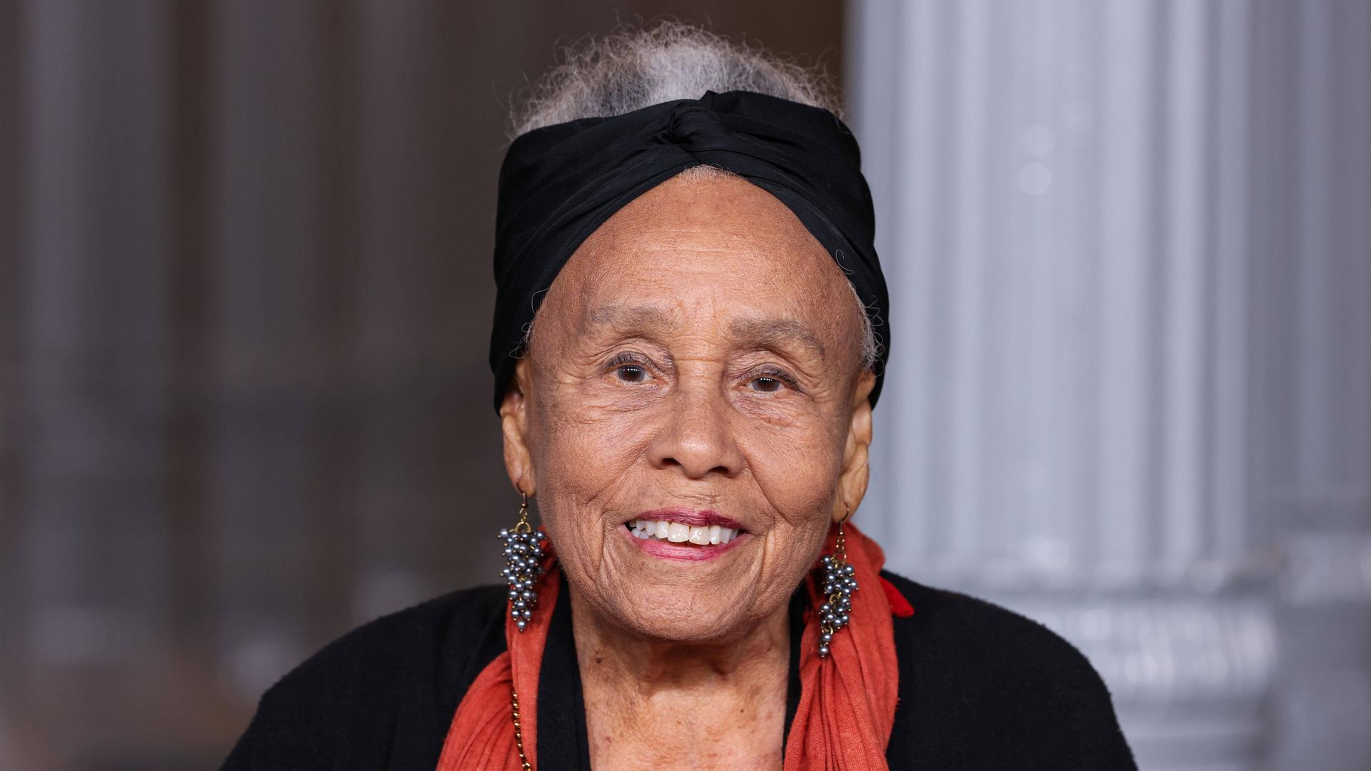 Betye Saar trägt ein schwarzes Kostüm, einen roten Schal und ein schwarzes Stirnband und lächelt am 1. November 2025 in die Kamera 