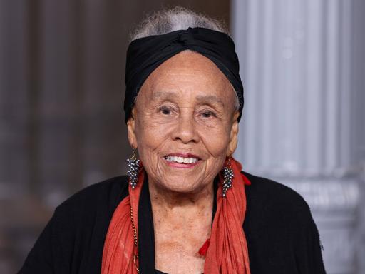 Betye Saar trägt ein schwarzes Kostüm, einen roten Schal und ein schwarzes Stirnband und lächelt am 1. November 2025 in die Kamera 