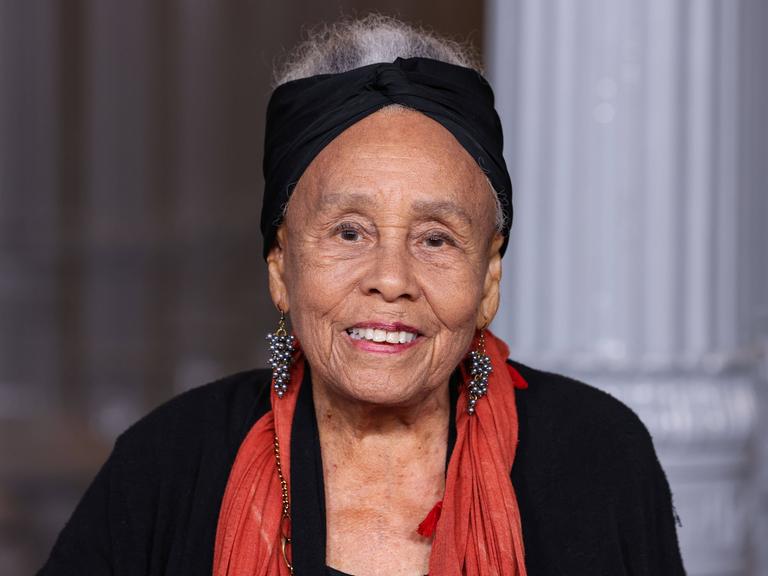 Betye Saar trägt ein schwarzes Kostüm, einen roten Schal und ein schwarzes Stirnband und lächelt am 1. November 2025 in die Kamera 