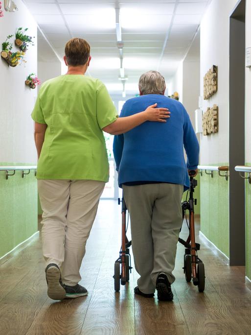 Eine Pflegefachkraft geht mit einer Bewohnerin durch ein Seniorenheim. Sie legt ihre Hand auf den Rücken der Frau. 