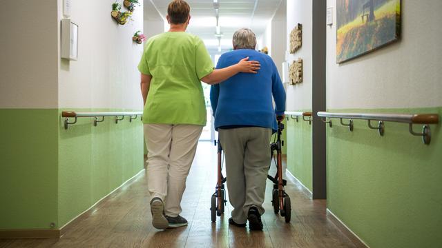 Eine Pflegerin geht mit einer Bewohnerin durch ein Senioren-Heim. Die Bewohnerin schiebt einen Rollator. Die Hand von der Pflegerin liegt auf dem Rücken von der Bewohnerin. 