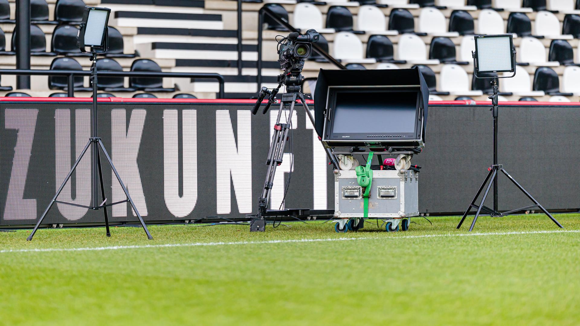 Kamera, TV und Monitor stehen am Rand eines Bundesligastadions. Im Hintergrund sind die Tribünen des Stadions zu sehen. 