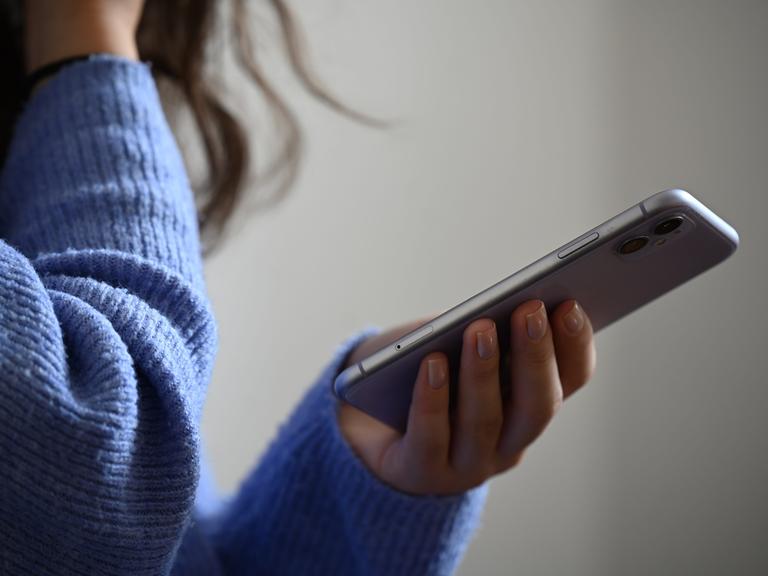 Eine Frau hält ein Smartphone in der Hand. Sie trägt einen blauen kuscheligen Wollpullover.