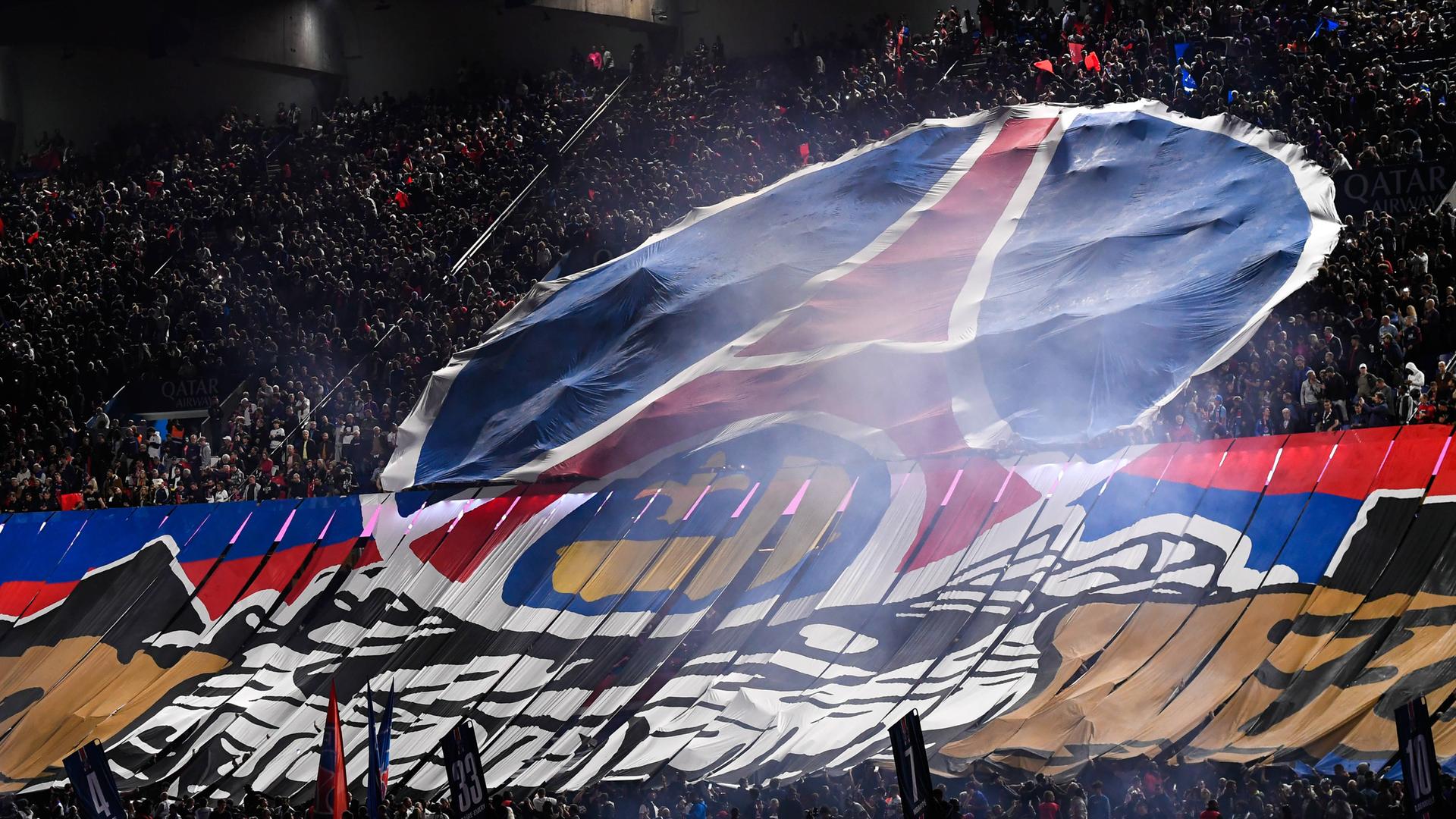 Das Bild zeigt eine Tribüne im Pariser Prinzenparkstadion. Fans haben eine Stofftransparent mit dem Logo des Vereins Paris Saint-Germain entfaltet.