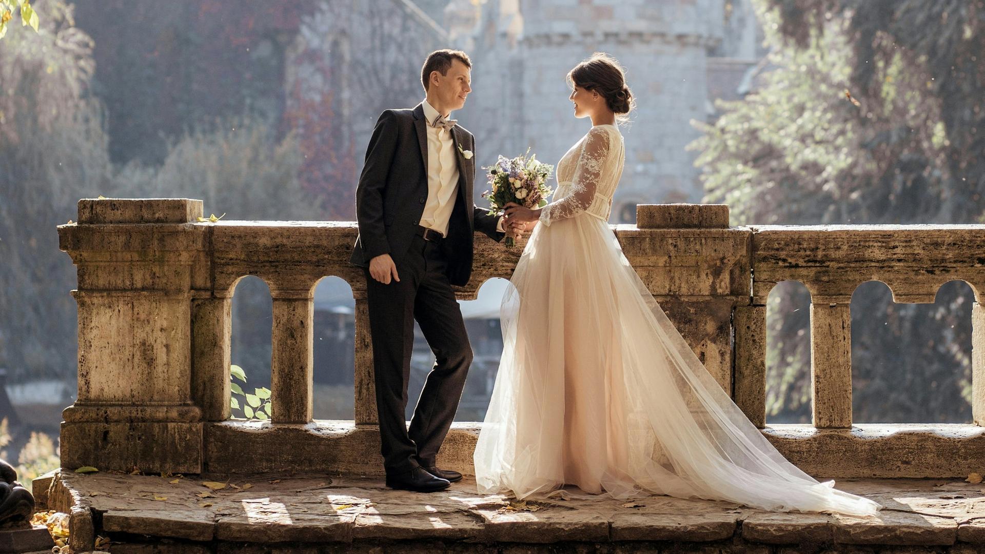 Wir sehen ein Hochzeitspaar vor einem Schloss. 