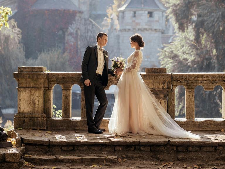 Wir sehen ein Hochzeitspaar vor einem Schloss. 
