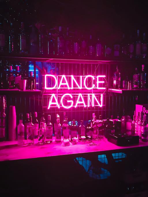 Hinter einer Bar ist in pinkfarbener Neonschrift der Schriftzug "DANCE AGAIN" angebracht.