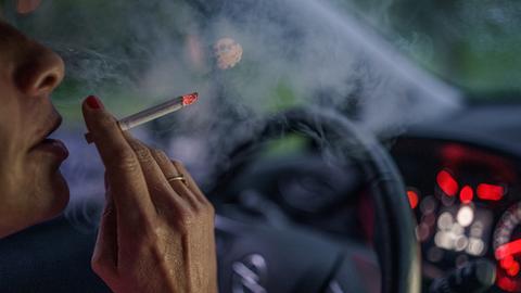 Eine Frau sitzt mit einer brennenden Zigarette hinter dem Lenkrad ihres Autos.