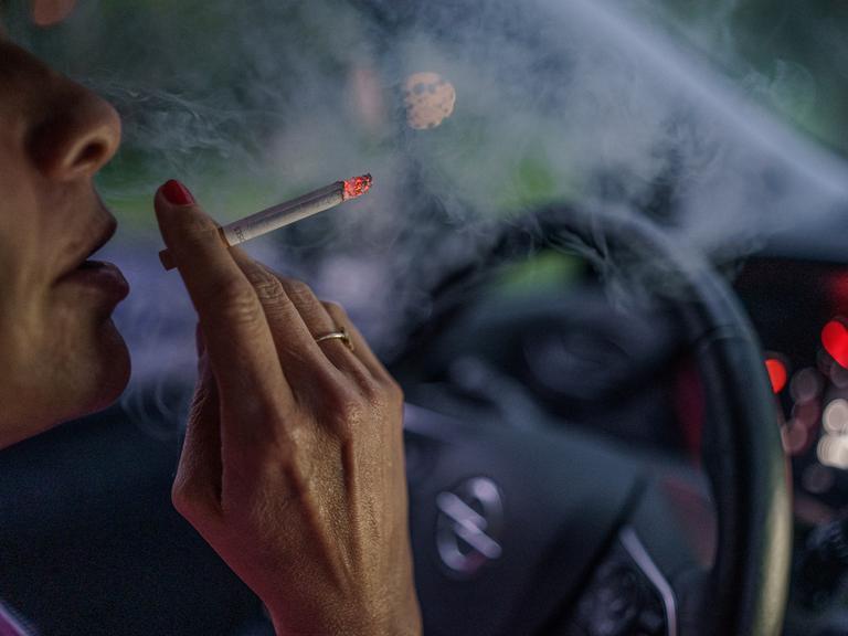 Eine Frau sitzt mit einer brennenden Zigarette hinter dem Lenkrad ihres Autos.