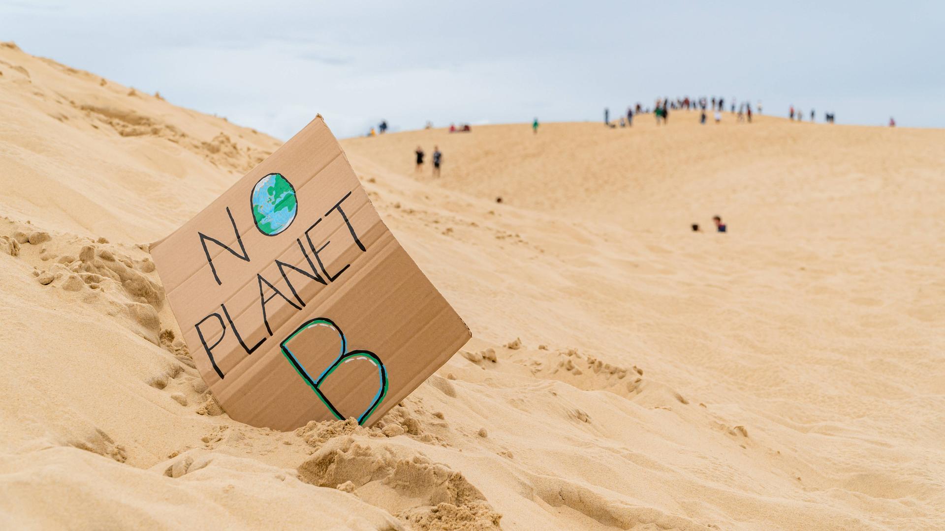 Eine Sanddünenlandschaft mit einem handgefertigten Schild mit der Aufschrift „No Planet B“ mit nicht erkennbaren Touristen auf Grande Dune du Pilat in Frankreich im Hintergrund. Eine Sanddünenlandschaft mit einem handgefertigten Schild mit der Aufschrift „No Planet B“ mit nicht erkennbaren Touristen auf Grande Dune du Pilat in Frankreich im Hintergrund.