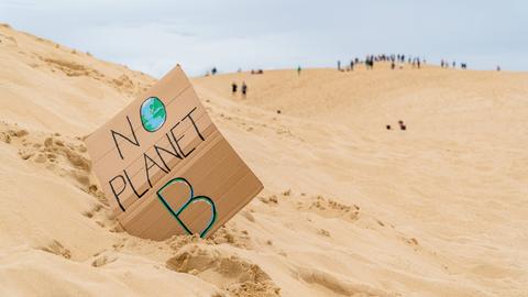 Eine Sanddünenlandschaft mit einem handgefertigten Schild mit der Aufschrift „No Planet B“ mit nicht erkennbaren Touristen auf Grande Dune du Pilat in Frankreich im Hintergrund. 