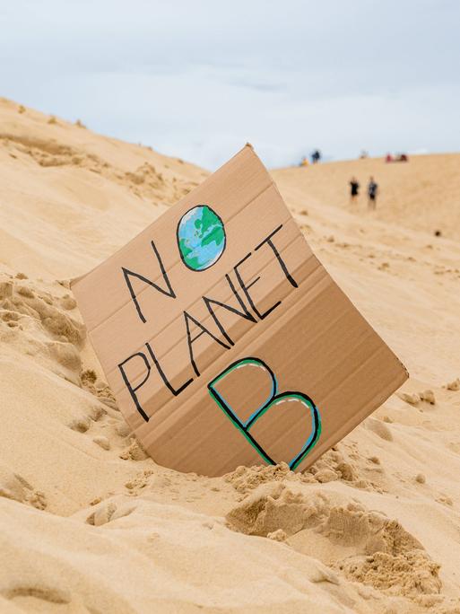 Eine Sanddünenlandschaft mit einem handgefertigten Schild mit der Aufschrift „No Planet B“ mit nicht erkennbaren Touristen auf Grande Dune du Pilat in Frankreich im Hintergrund. 