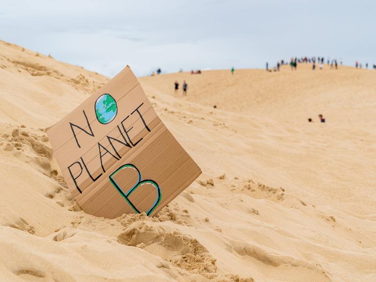 Eine Sanddünenlandschaft mit einem handgefertigten Schild mit der Aufschrift „No Planet B“ mit nicht erkennbaren Touristen auf Grande Dune du Pilat in Frankreich im Hintergrund. 