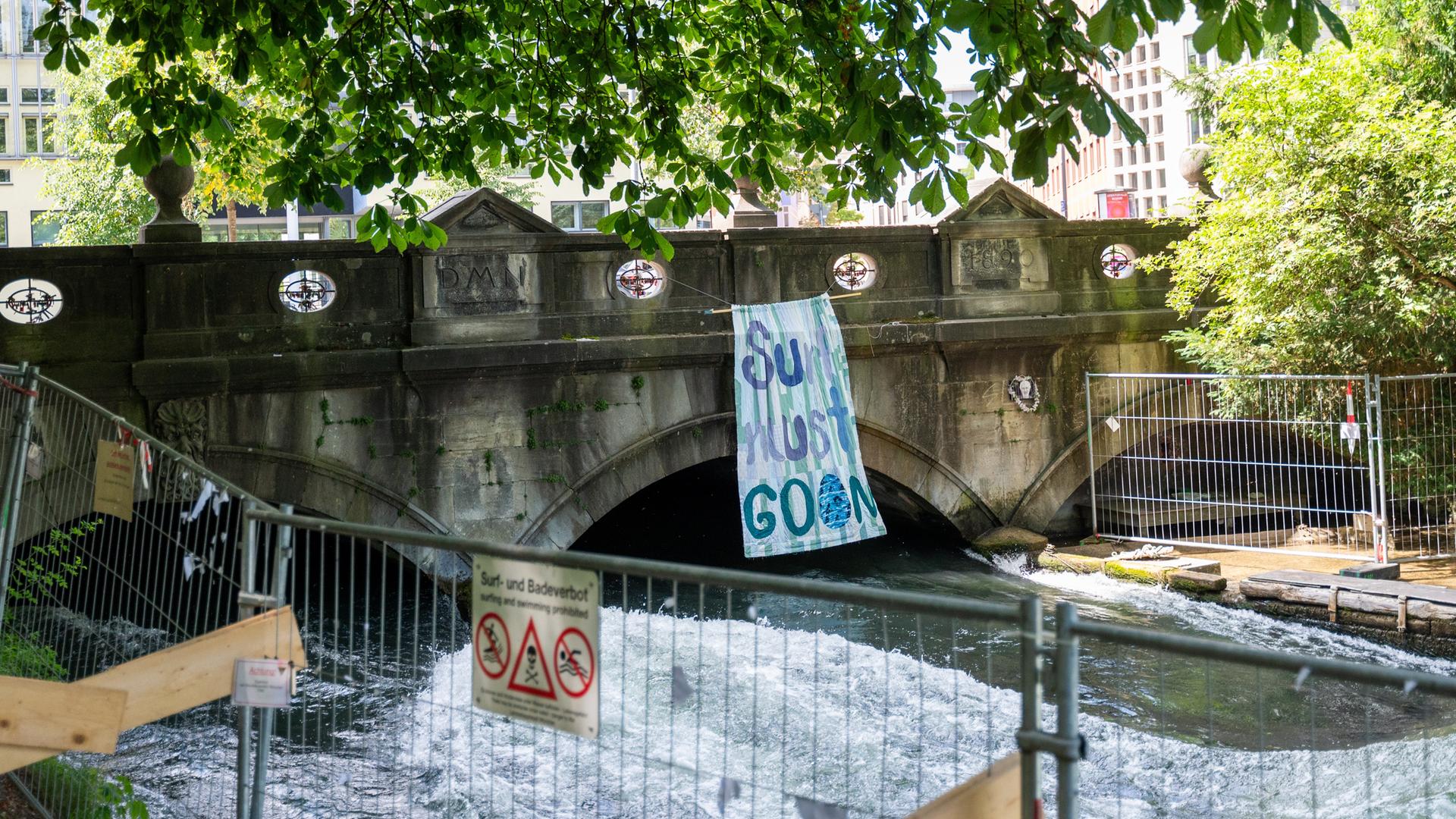 München: Ein Banner mit der Aufschrift "Surf must go on" hängt an der Eisbachbrücke an der Eisbachwelle. Im Vordergrund sieht man Absperrungen. München: Ein Banner mit der Aufschrift "Surf must go on" hängt an der Eisbachbrücke an der Eisbachwelle. Im Vordergrund sieht man Absperrungen.