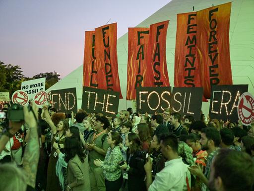 Menschen halten Schilder mit der Aufschrift "End The Fossil Era" während eines Protests. Beim Endspurt der Weltklimakonferenz gibt es nun Bewegung..