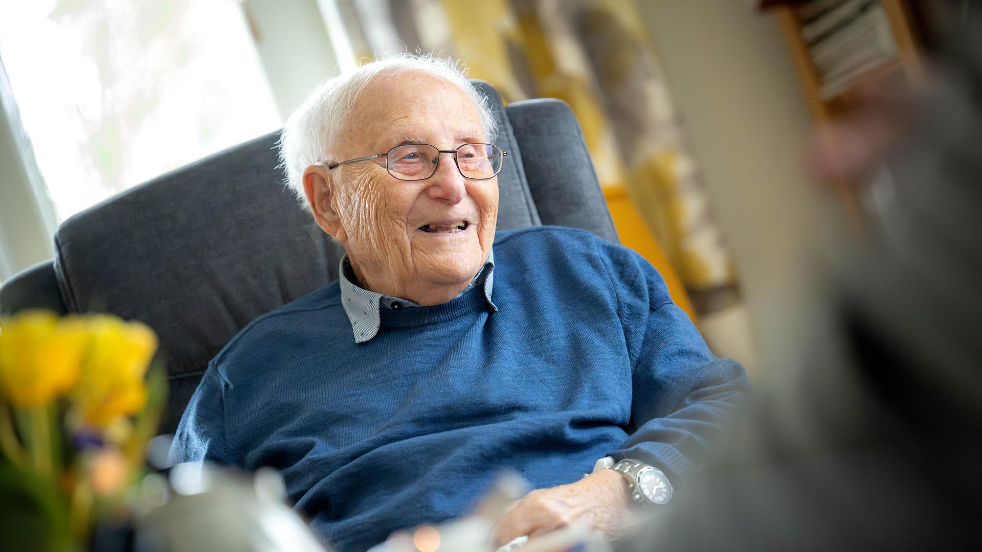 Albrecht Weinberg sitzt in seiner Wohnung in Ostfriesland. Er trägt ein graues Hemd, darüber einen blauen Pullover, eine Brille und guckt in dei Kamera. Albrecht Weinberg sitzt in seiner Wohnung in Ostfriesland. Er trägt ein graues Hemd, darüber einen blauen Pullover, eine Brille und guckt in dei Kamera.