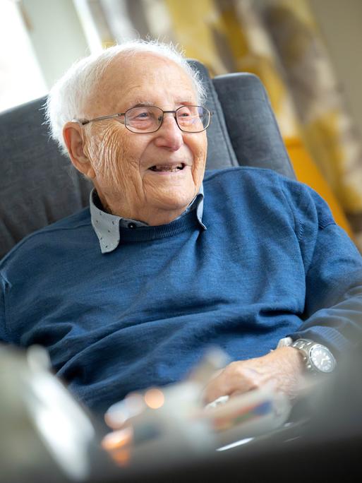 Albrecht Weinberg sitzt in seiner Wohnung in Ostfriesland. Er trägt ein graues Hemd, darüber einen blauen Pullover, eine Brille und guckt in dei Kamera.