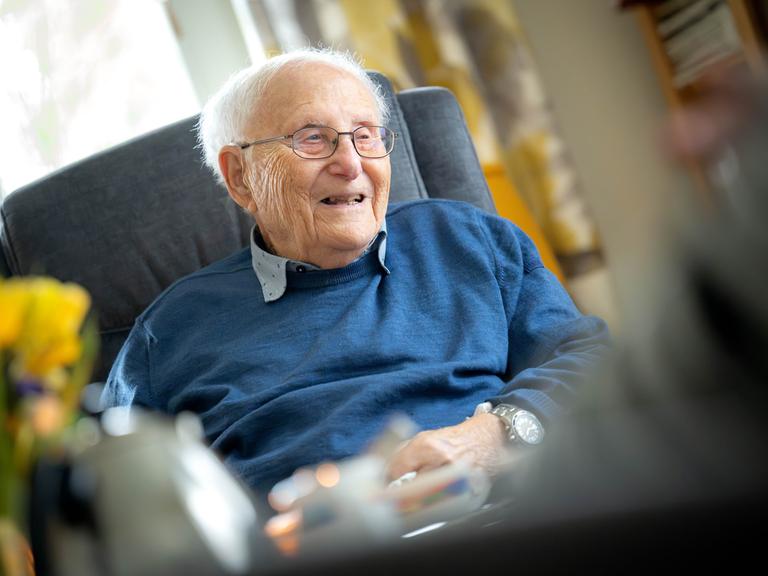 Albrecht Weinberg sitzt in seiner Wohnung in Ostfriesland. Er trägt ein graues Hemd, darüber einen blauen Pullover, eine Brille und guckt in dei Kamera.
