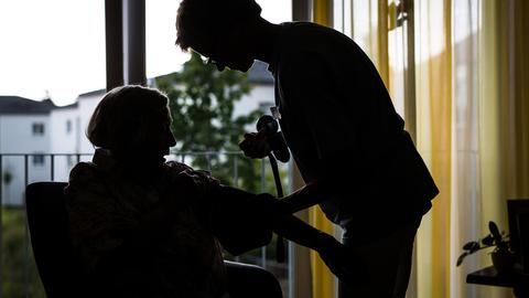 Eine Pflegekraft kümmert sich in einem Pflegeheim um eine ältere Dame und kontrolliert den Blutdruck. Das Bild wirkt etwas düster.