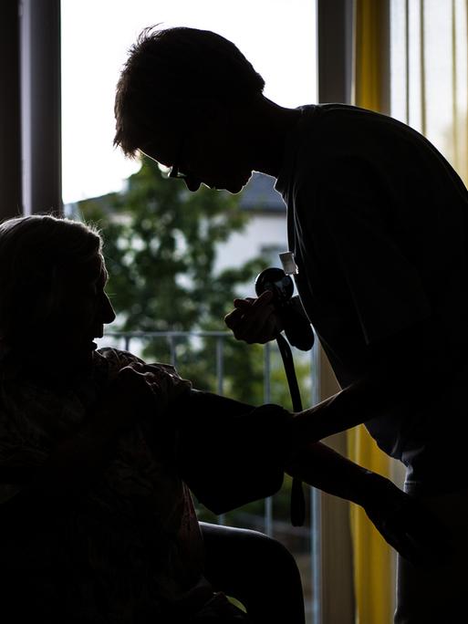 Eine Pflegekraft kümmert sich in einem Pflegeheim um eine ältere Dame und kontrolliert den Blutdruck. Das Bild wirkt etwas düster.