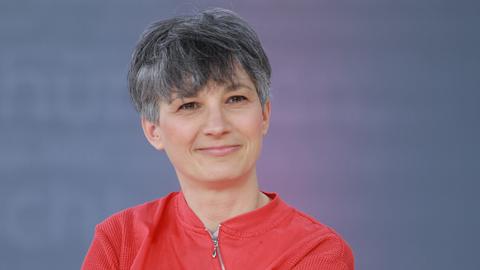 Stefanie de Velasco sitzt auf einer Bühne bei der Leipziger Buchmesse vor einem blauen Hintergrund. Sie trägt eine rote Jacke und lächelt in die Kamera. Stefanie de Velasco sitzt auf einer Bühne bei der Leipziger Buchmesse vor einem blauen Hintergrund. Sie trägt eine rote Jacke und lächelt in die Kamera.