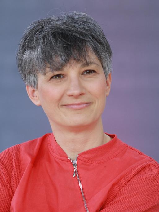 Stefanie de Velasco sitzt auf einer Bühne bei der Leipziger Buchmesse vor einem blauen Hintergrund. Sie trägt eine rote Jacke und lächelt in die Kamera.