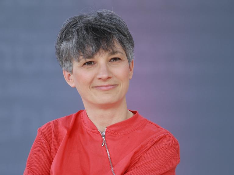 Stefanie de Velasco sitzt auf einer Bühne bei der Leipziger Buchmesse vor einem blauen Hintergrund. Sie trägt eine rote Jacke und lächelt in die Kamera.