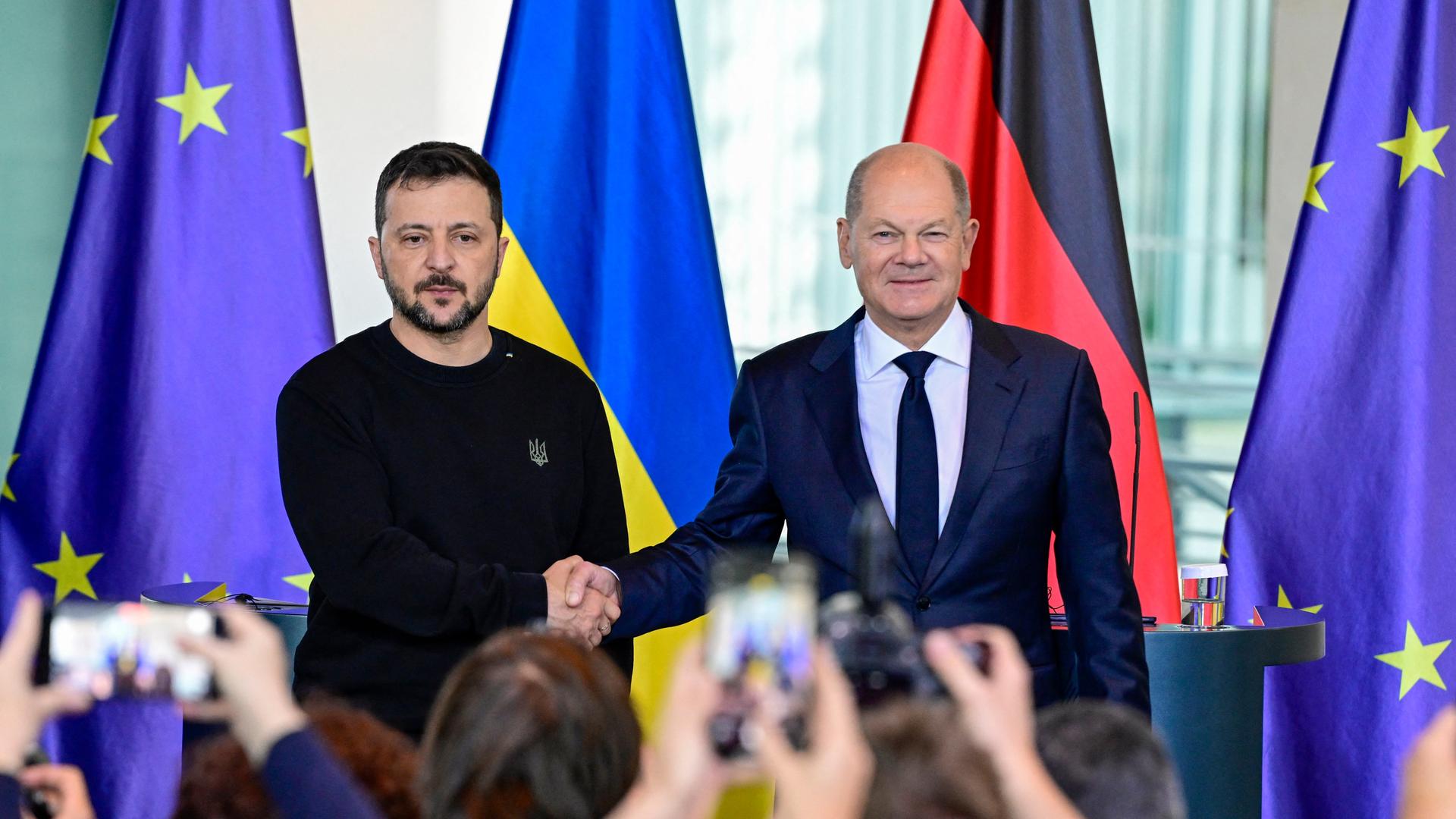 Das Bild zeigt den ukrainischen Präsidenten Selenskyj mit Bundeskanzler Scholz bei einer Pressekonferenz. Sie geben sich die Hände. 